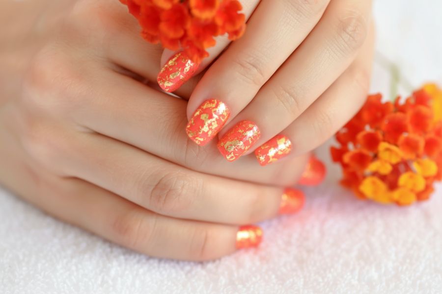 Bright coral square nails with gold foil accents, complemented by matching orange flowers.