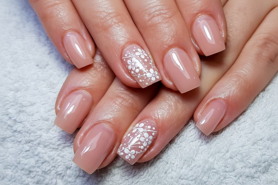 Glossy nude square nails with white floral designs on accent fingers over a soft towel background.