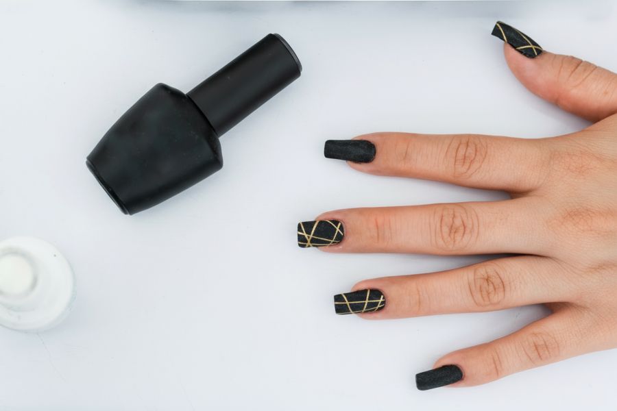 Hand with matte black square nails featuring gold geometric line designs on a white background.