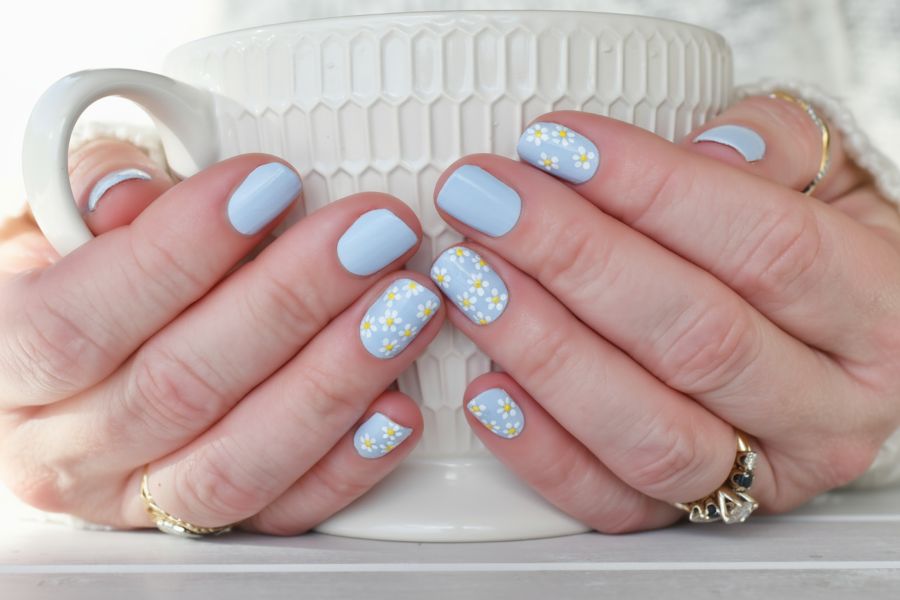 Hands with pastel blue square nails featuring small white daisies, holding a white textured mug.