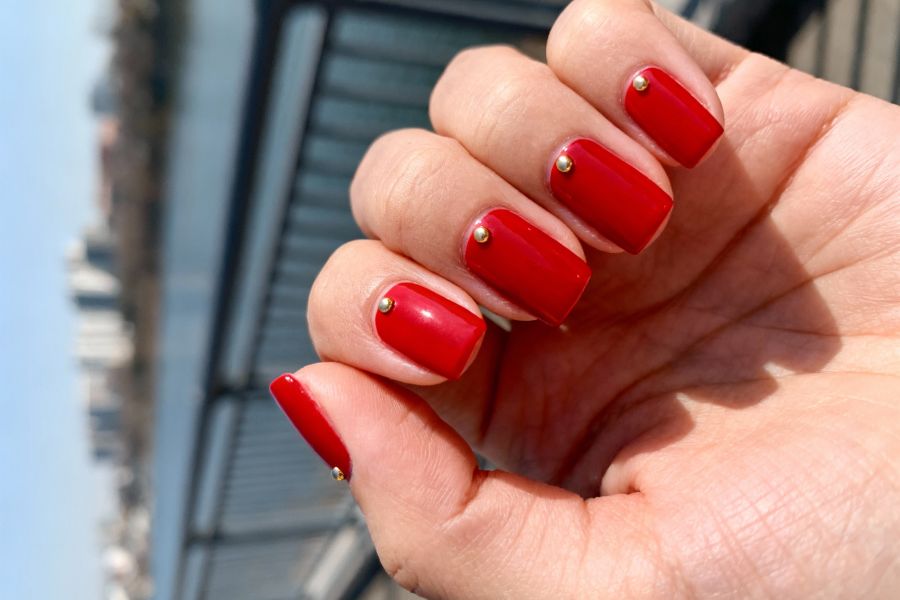 Close-up of shiny red square nails accented with small gold studs, captured outdoors by the water.