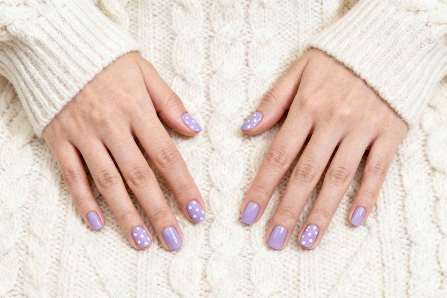 Hands with soft lavender square nails featuring small white polka dots on a cream knit sweater.