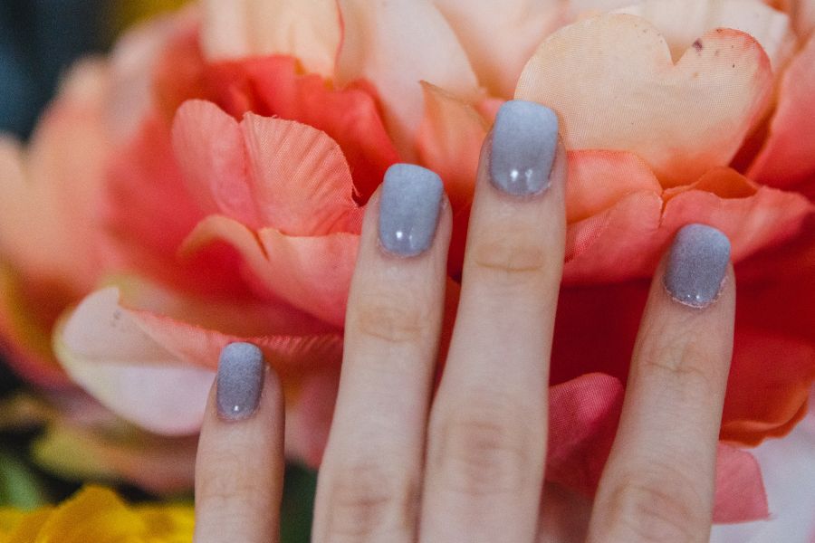 Hand with sparkly silver square nails featuring a soft gradient ombre design near pink petals.
