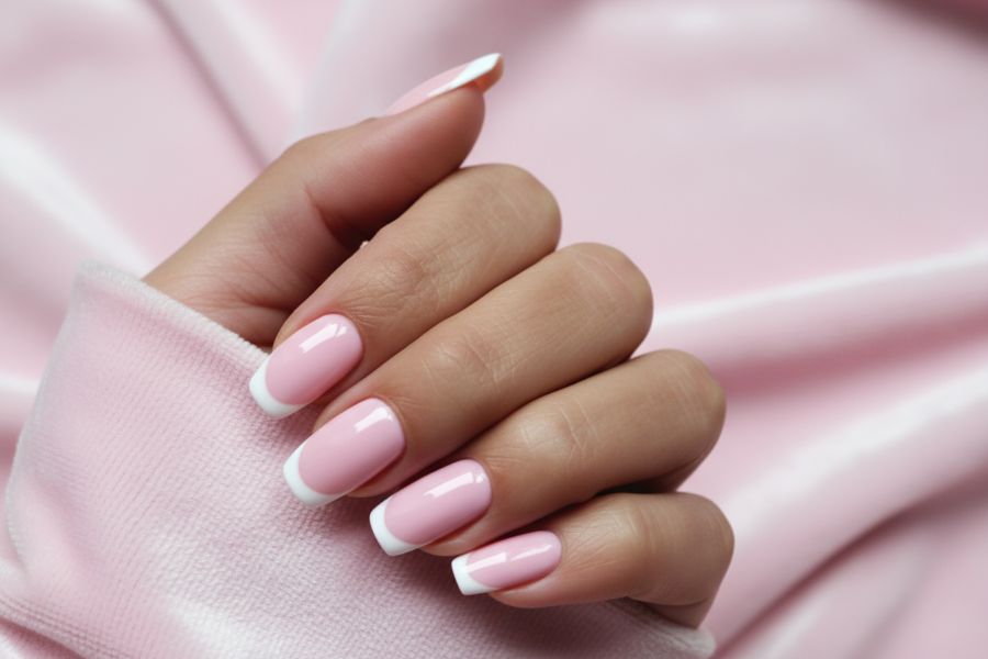 Close-up of pink square nails with thin white French tips on a soft pink fabric background.