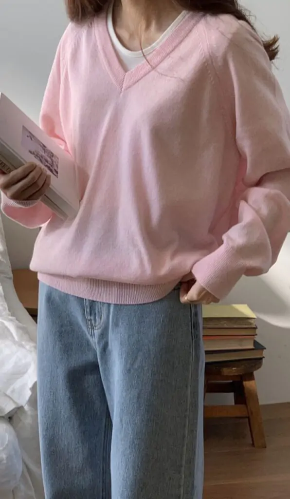 Woman in a pastel pink V-neck sweater with light-wash jeans holding a book, styled for a cozy fall day.