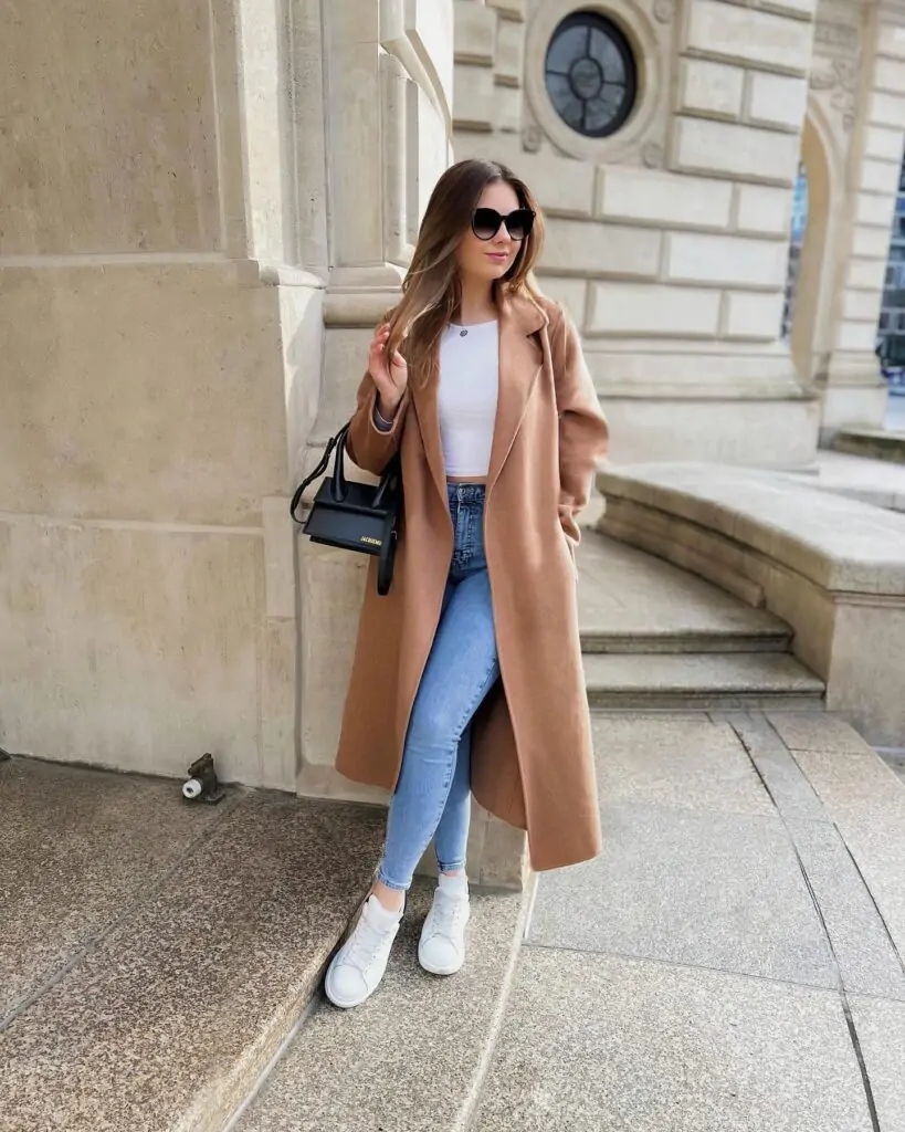 Fashionable person in classic fall outfit with camel coat, white top, blue jeans, white sneakers, and black shoulder bag.