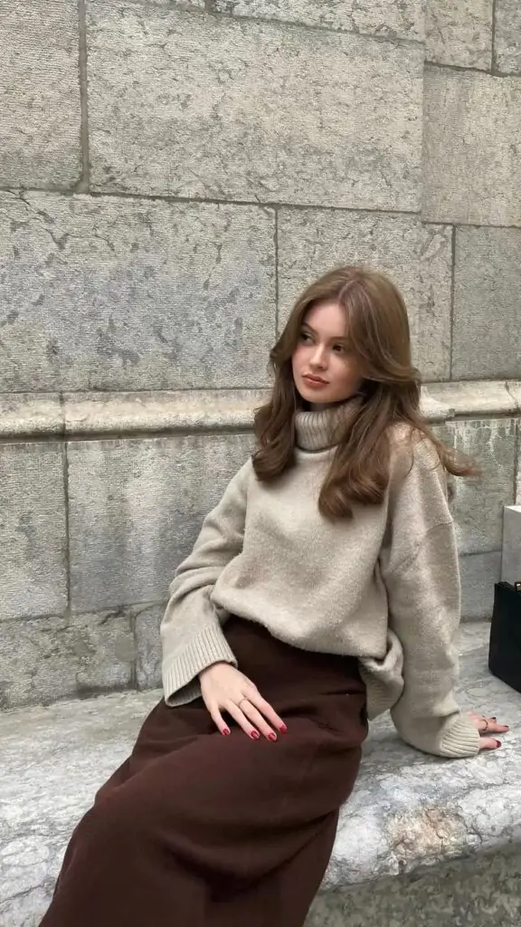 Woman in a beige turtleneck sweater and dark brown skirt sitting on a stone bench, showing a cozy fall outfit.