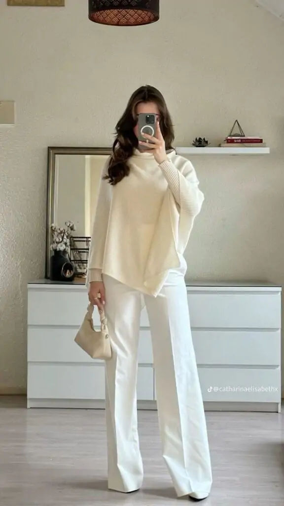 Woman in oversized cream poncho sweater with white wide-leg trousers and beige top-handle bag.