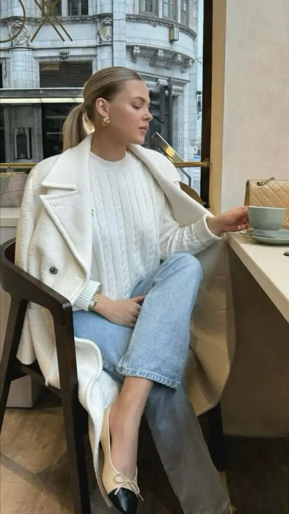 Woman in a white wool coat, cable-knit sweater, and light-wash jeans, enjoying coffee at a café.