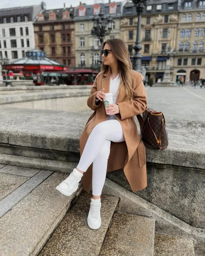 Fashionable individual in fall attire with camel coat and white jeans sitting on city steps.