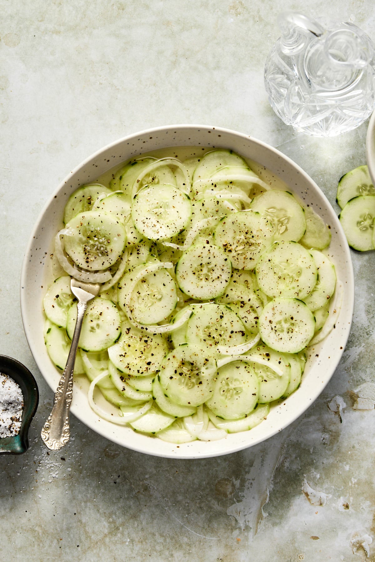 An overhead shot of cucumber vinegar salad in a white bowl with sliced onions and black pepper.