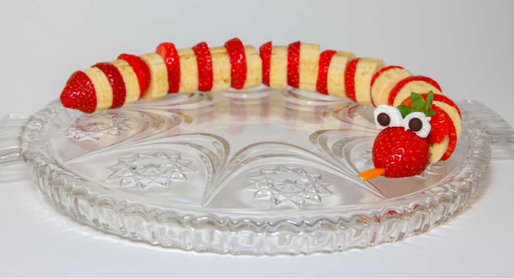 Strawberries and bananas combined with mini marshmallows and chocolate chips to make a snake sigging on a glass dish.