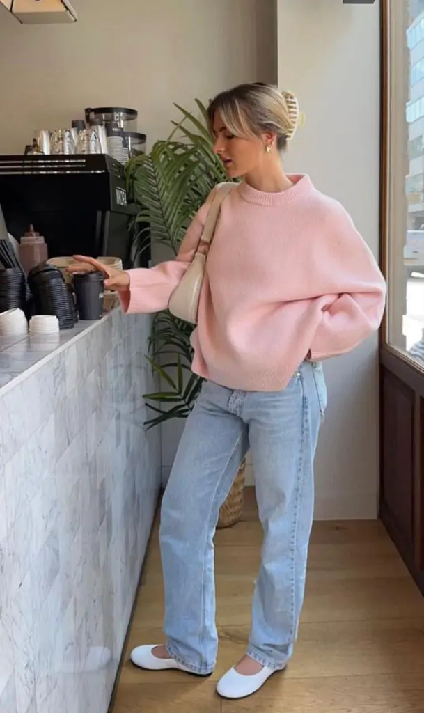 Woman in an oversized pink sweater with light wash jeans and white ballet flats, holding a neutral shoulder bag at a café counter.