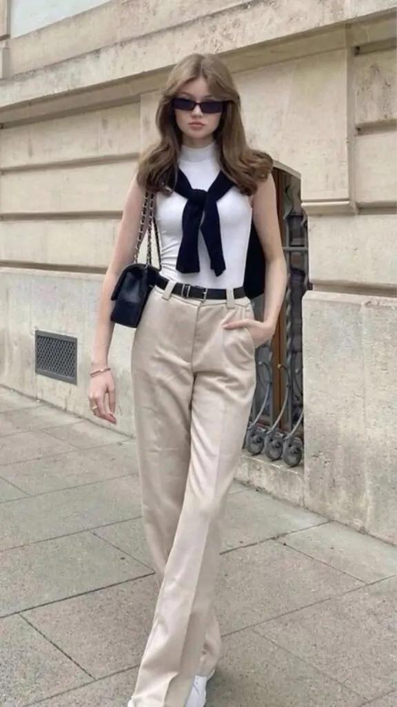 Fashionable person in fall attire with white top, black bow tie, beige trousers, black shoulder bag, and white sneakers against an urban backdrop.
