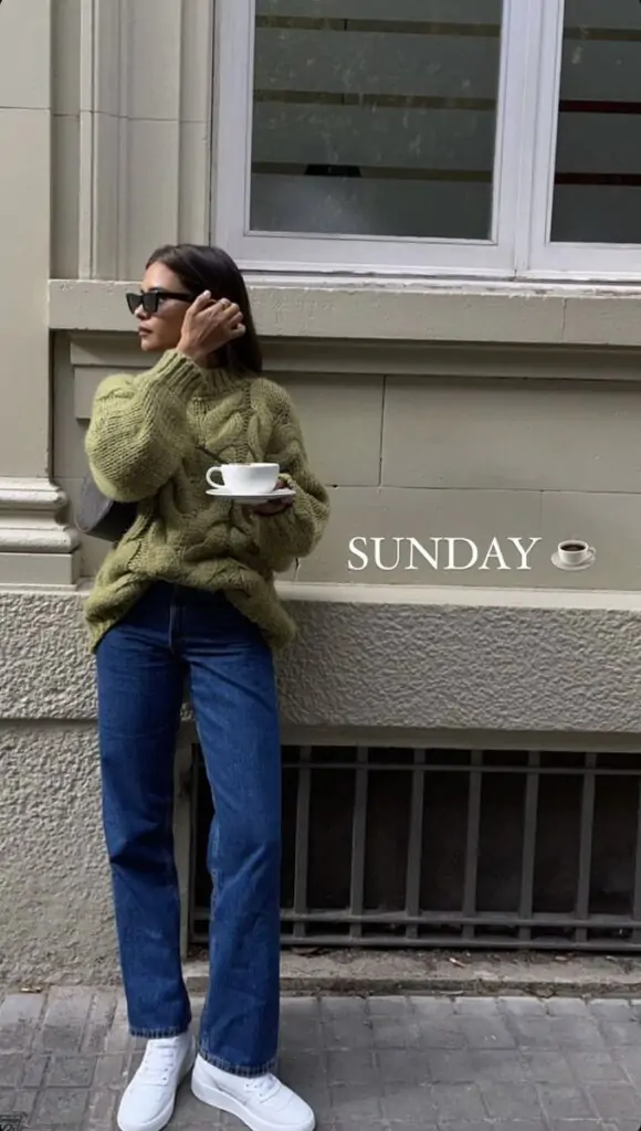 Woman wearing an olive chunky knit sweater, blue jeans, white sneakers, and black sunglasses while holding a coffee cup, standing against a building.