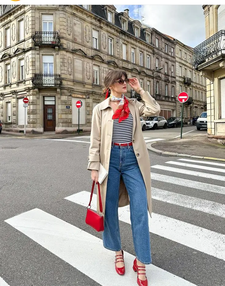 Fashionable individual crossing street in chic fall outfit featuring beige trench coat, striped shirt, blue jeans, red scarf, handbag, and sandals.