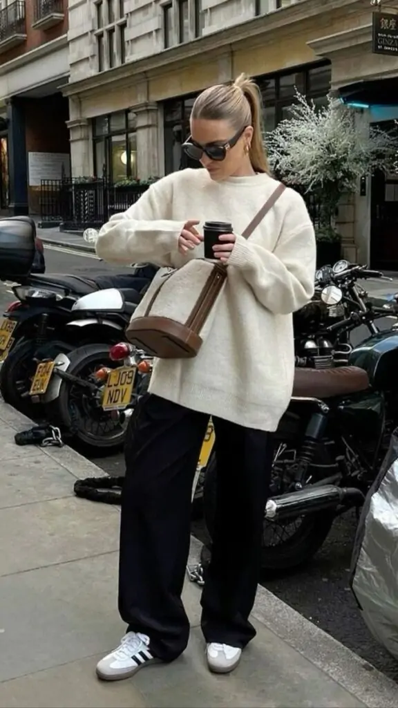 Stylish woman in a chunky cream sweater and black trousers on a fall day in the city, accessorized with a crossbody bag and classic sneakers.