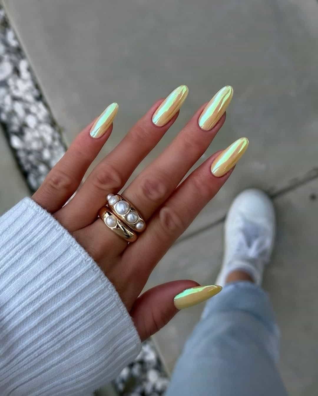 long almond shaped iridescent yellow chrome nails on a hand wearing gold pearl rings